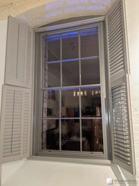 Colour matched magnetic secondary glazing with split panel and subframe, fitted to a grey sash window with grey plantation shutters